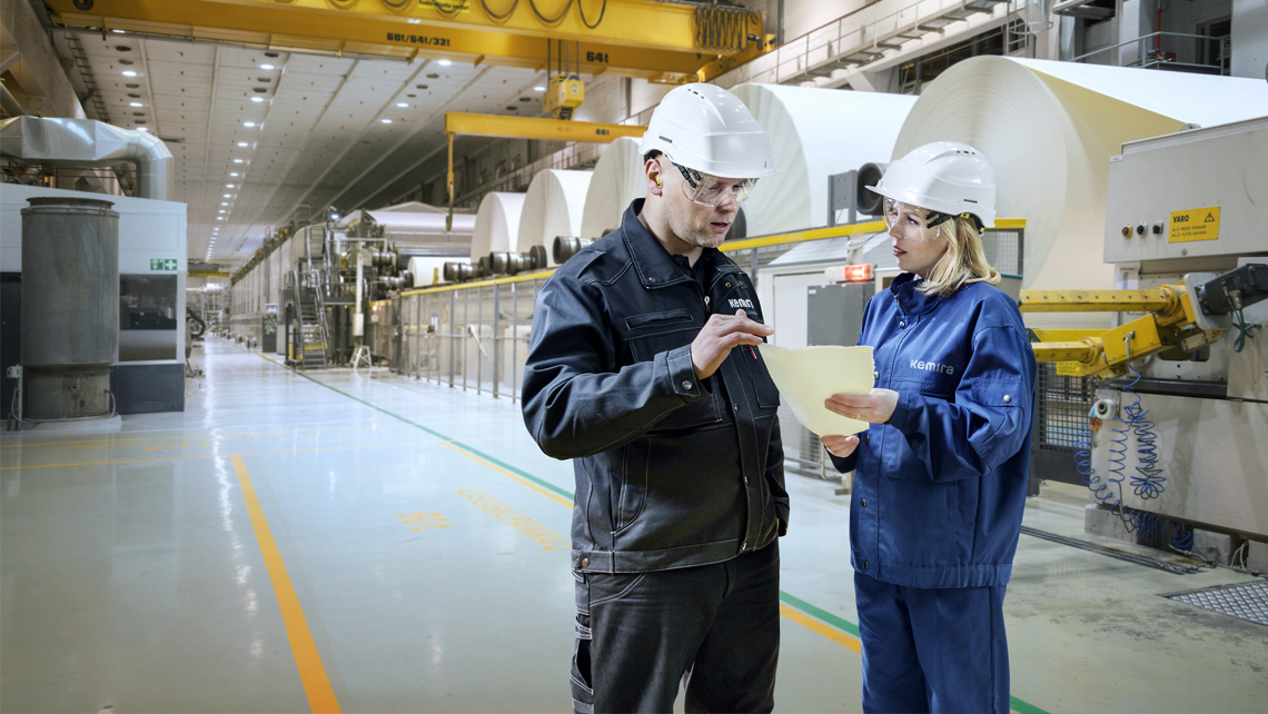 Two workers standing in the front of paper machine rolls.