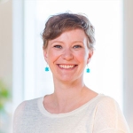 Woman with short brown hair white long sleeve shirt and blue earrings smiling at the camera.