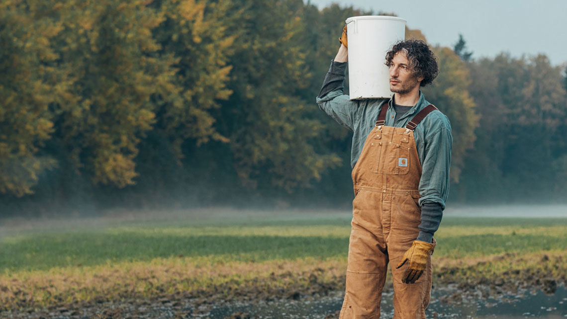 Farmer holding white bucket standing in muddy fields.