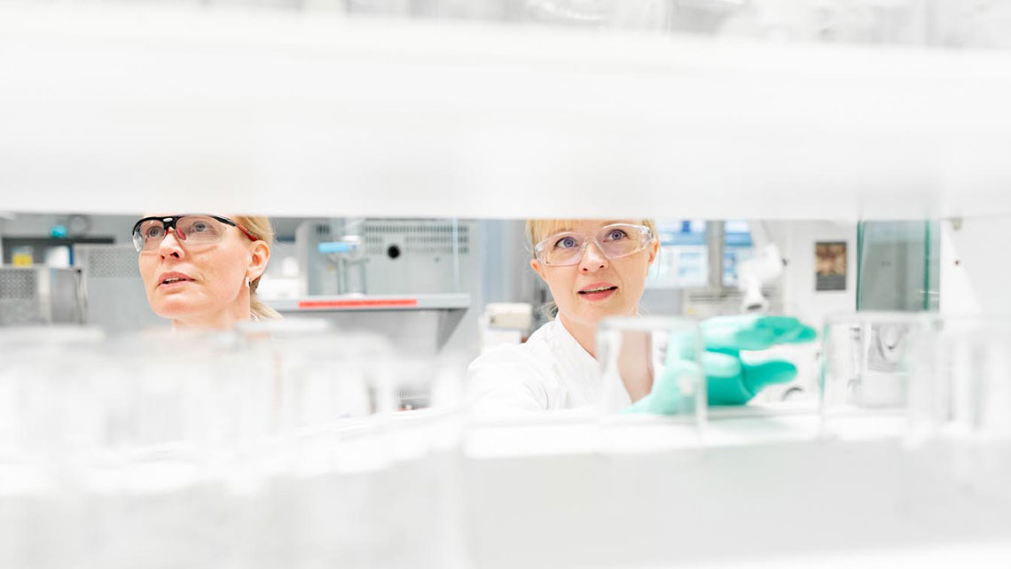 Two persons in a laboratory working together with blurry white background and blurred lab equipment.