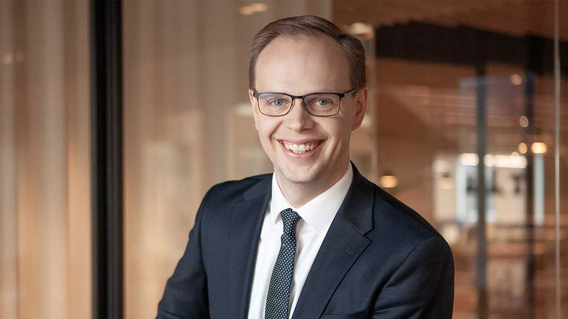 young man in blue business suit with white shirt and blue tie smiling towards the camera.