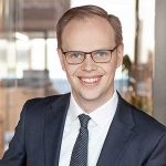 Young man in blue business suit with white shirt and blue tie smiling towards the camera.