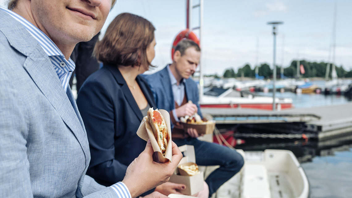 Three persons sitting in a harbour and eating takeway food.