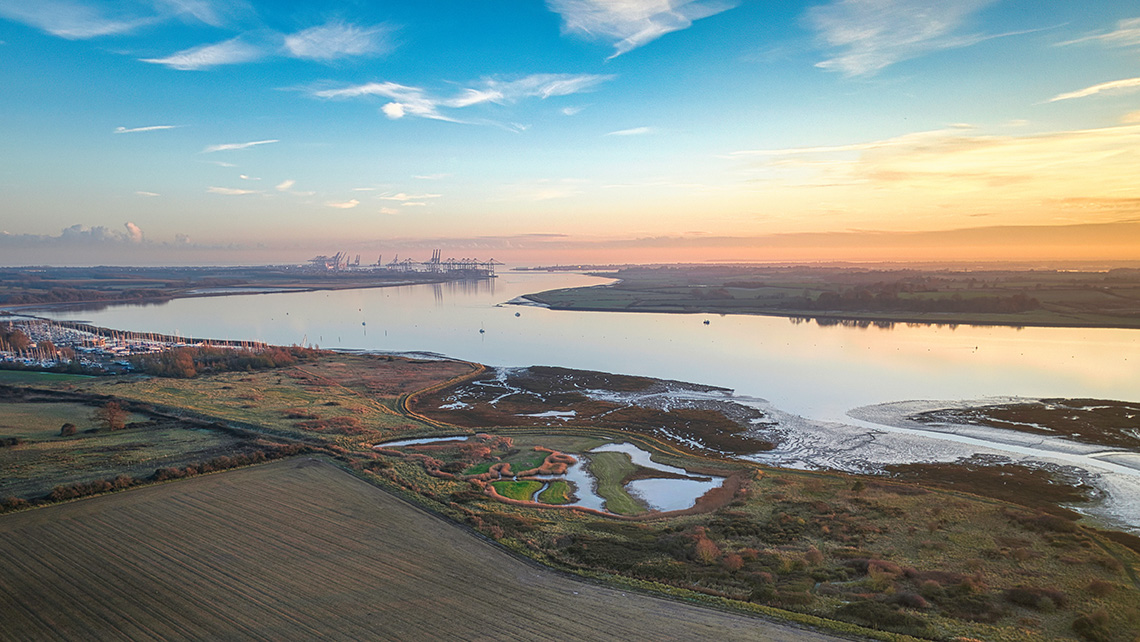 Paysage fluvial au coucher du soleil avec des champs et des zones humides au premier plan et des structures industrielles lointaines à l'horizon.