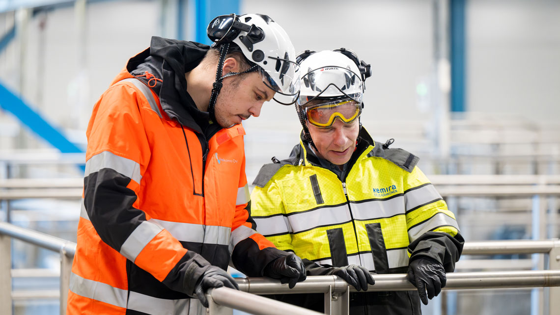 Two workers in safety gear, including helmets and high-visibility jackets, discussing something while leaning over a metal railing. They appear to be in an industrial or wastewater treatment facility.