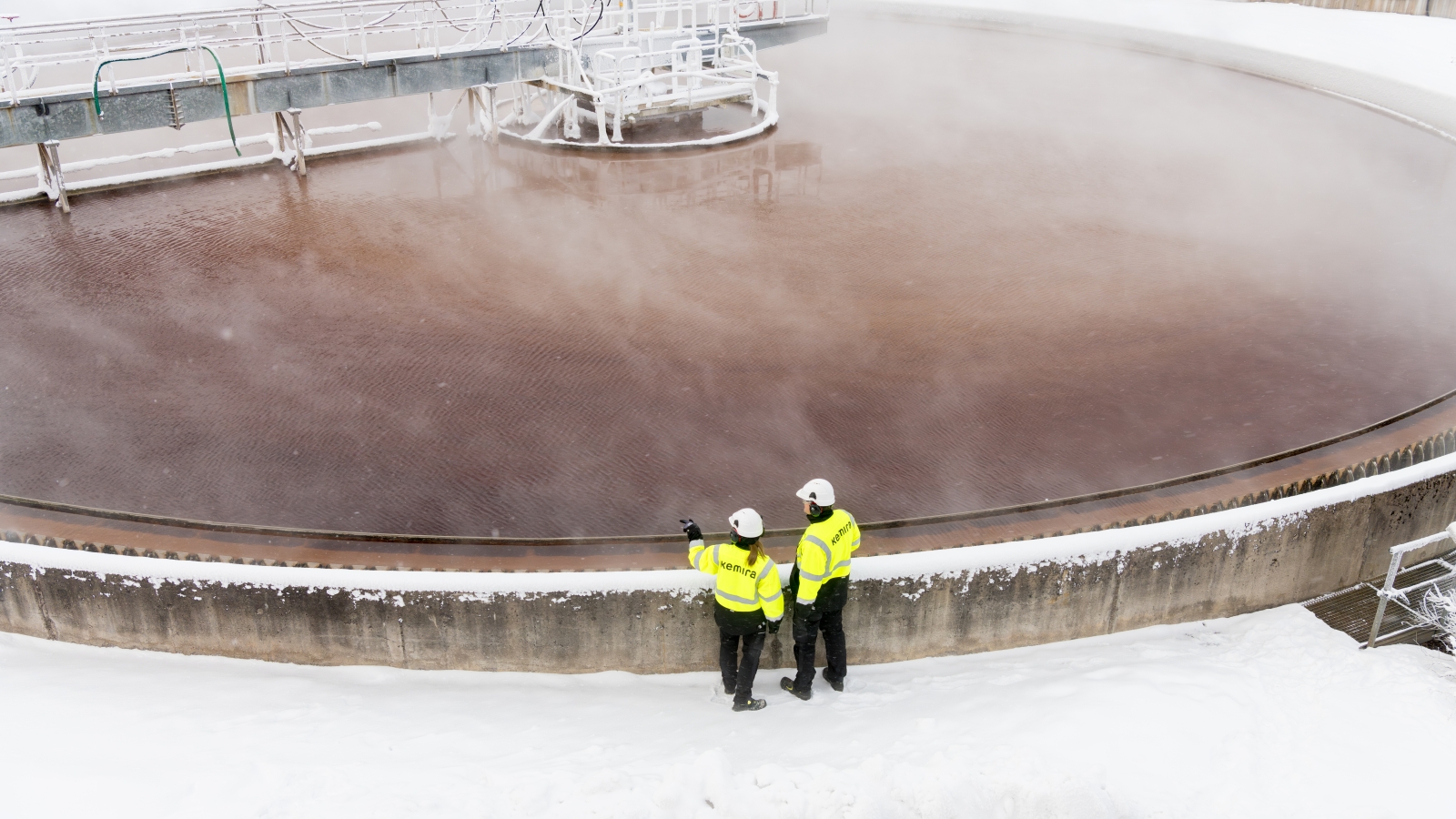 Two workers wearing PPE stand near a large, circular water treatment clarifier in a snowy environment.