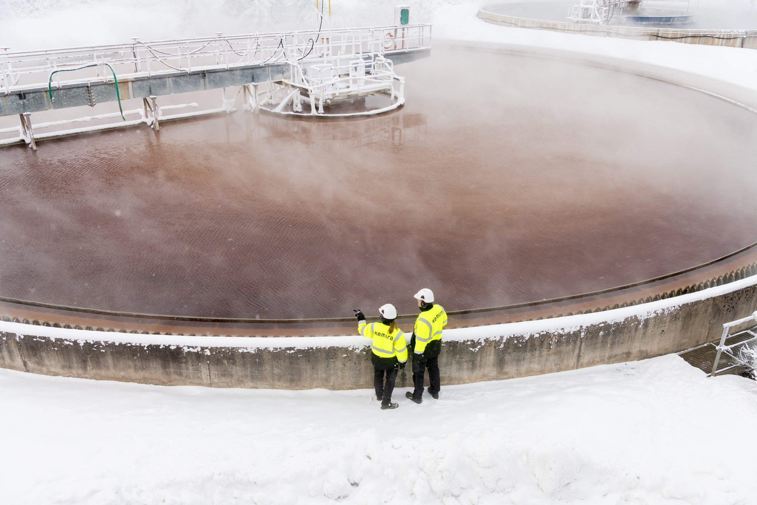 Two workers wearing PPE stand near a large, circular water treatment clarifier in a snowy environment.