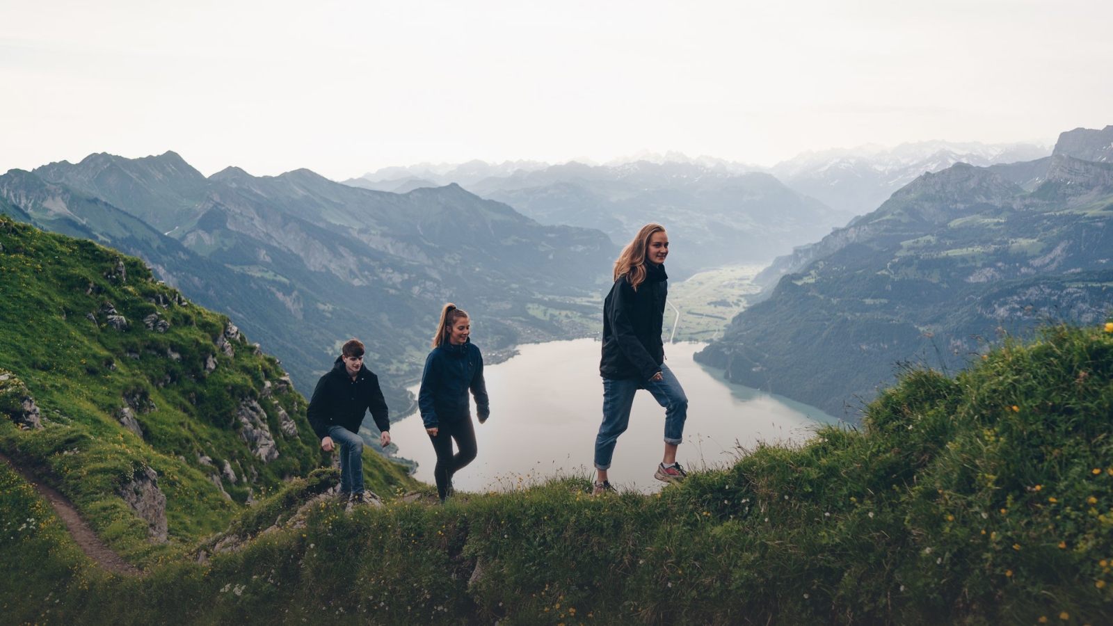 Grupo de amigos caminhando em trilha de montanha com lago cênico e picos alpinos ao fundo.