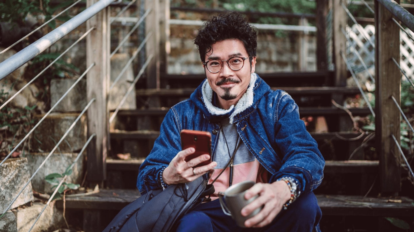 Young asian man smiling joyfully at camera while using smartphone in country side.